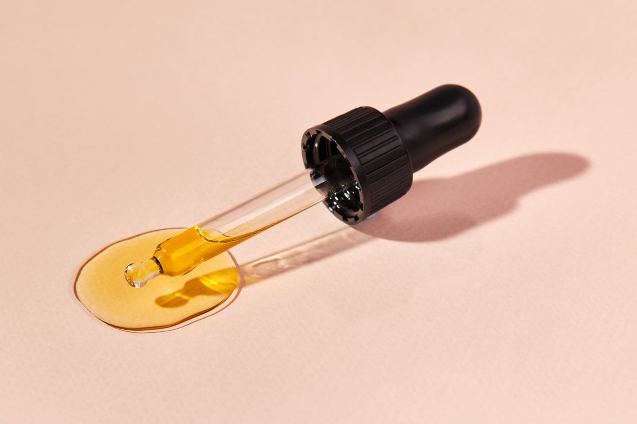 Breaking Down the Different Types of Retinoids 3 Close up of pipette with pouring liquid essential oil on beige background. Dropper with a falling drop of aromatherapy oil close-up