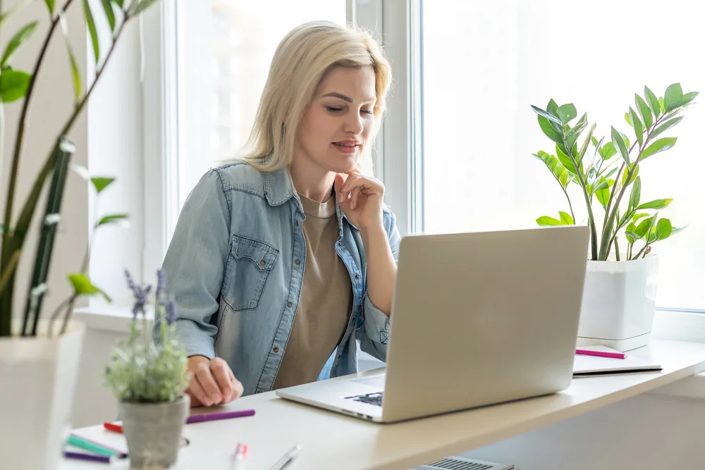 A woman surrounded by plants reading an article on her laptop titled odd ways to make money