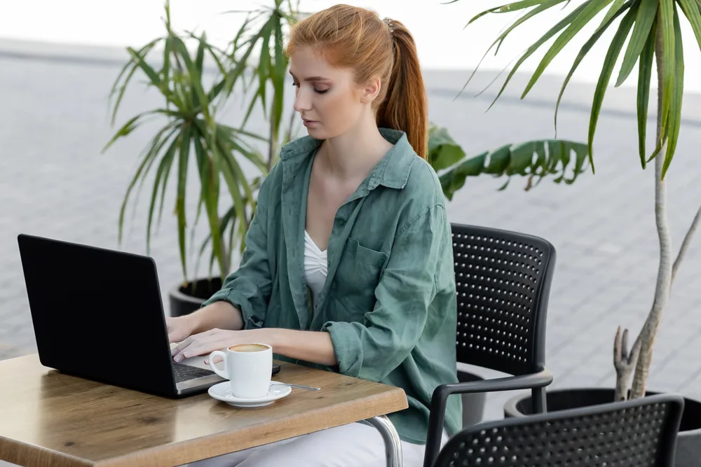 A young female with red hair using her laptop on a patio looking for online summer jobs for teachers