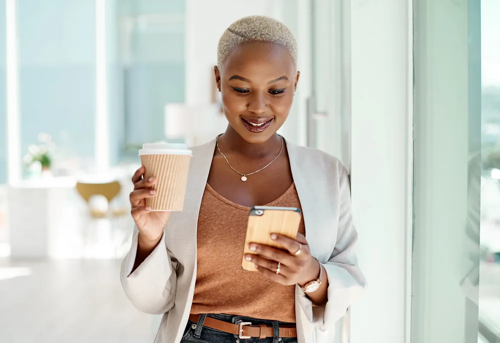 A young woman holding a to go coffee looking for weekend jobs on her mobile phone